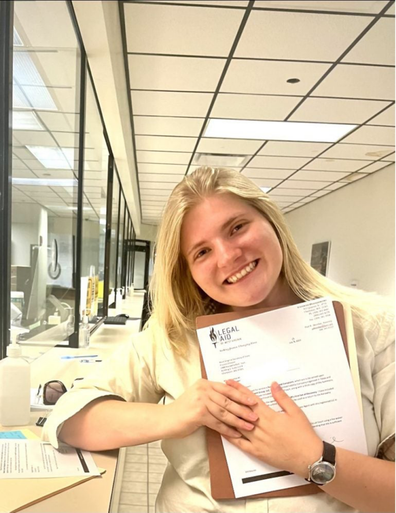 A blond college aged woman stands in a hallway holding a document and smiling.