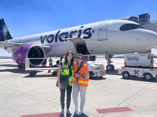 Two women in highlighted vests standing on an air strip in front of an airplane with the Volaris logo on the side.