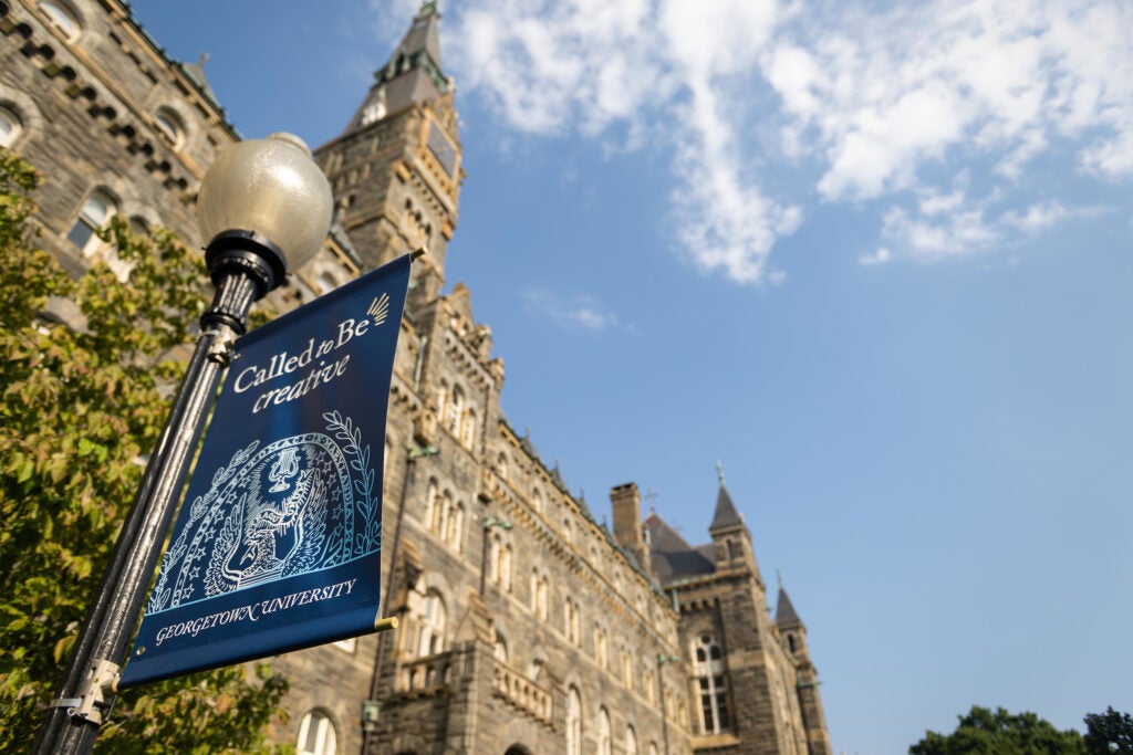 A lamppost with a navy poster reading "Called to Be Creative" in front of a stone building with a clock tower.