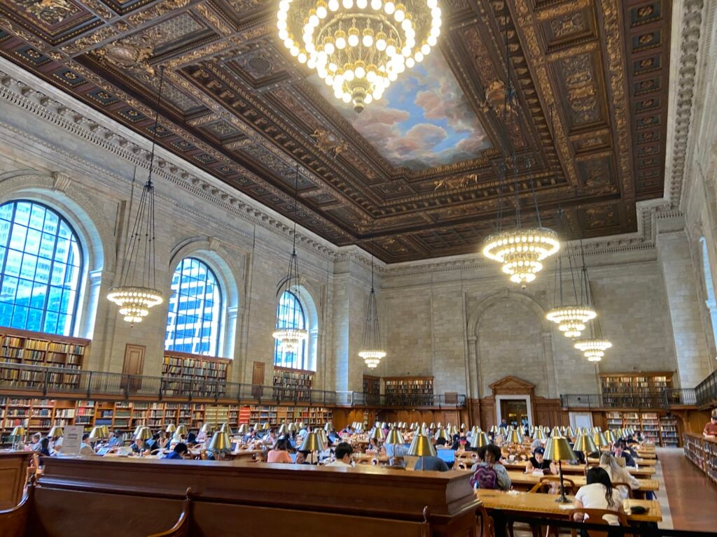 A large room with a painted ceiling, four chandeliers, and rows of long tables with people working at laptops.