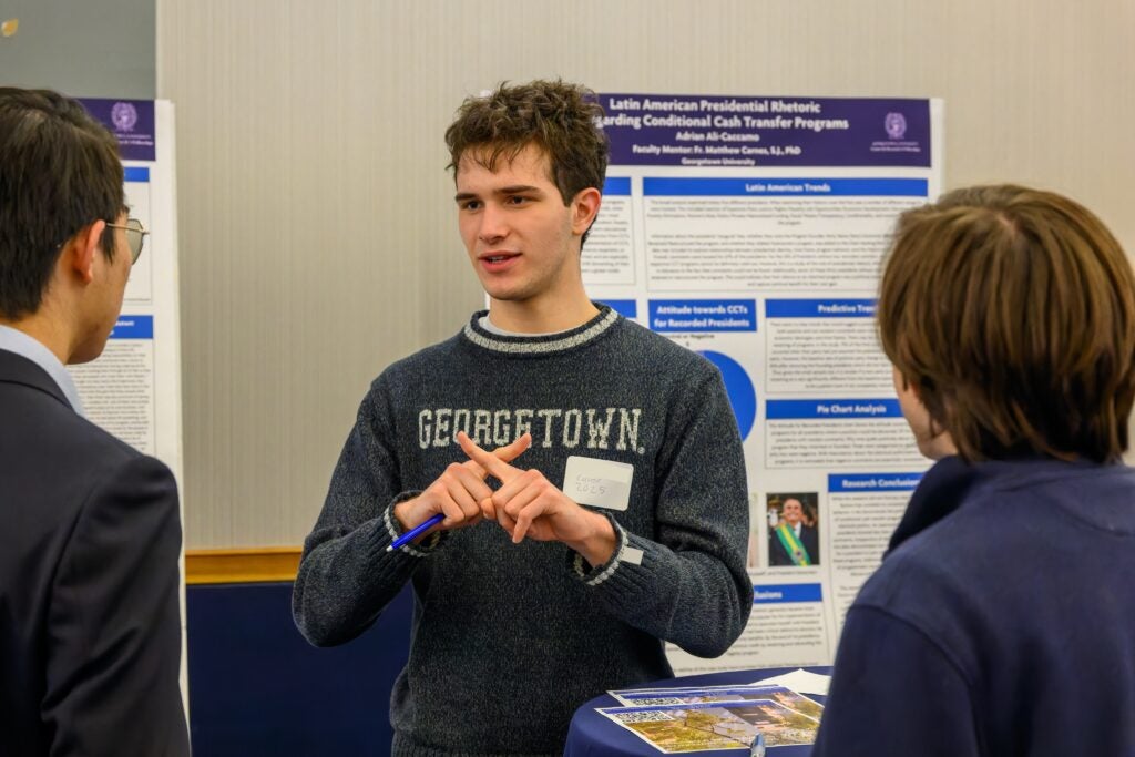 A student in a navy Georgetown sweater stands in front of a research poster speaking to two other students.