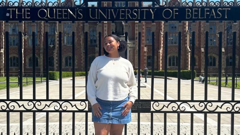 Mara Lewis stands in front of a gate that says: Queen's University Belfast with a building in the background