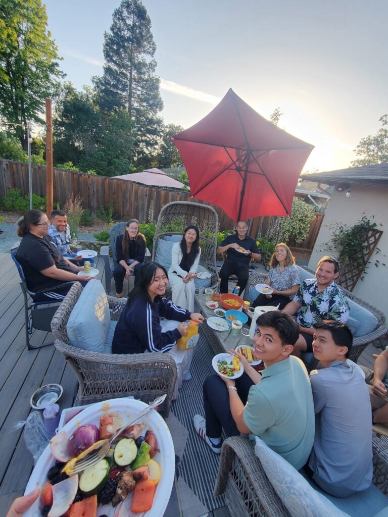 A group of Georgetown students and Livermore mentors sit in a circle on lawn chairs outside. Individuals are holding plates of food and facing towards middle table.