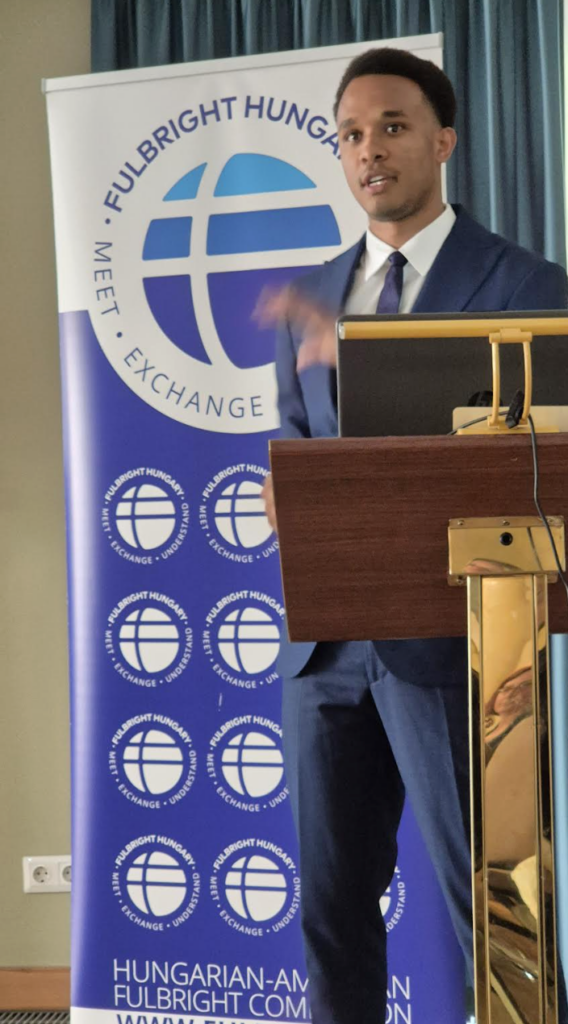 Noah Aire speaks at a podium in front of a Fulbright Hungary banner